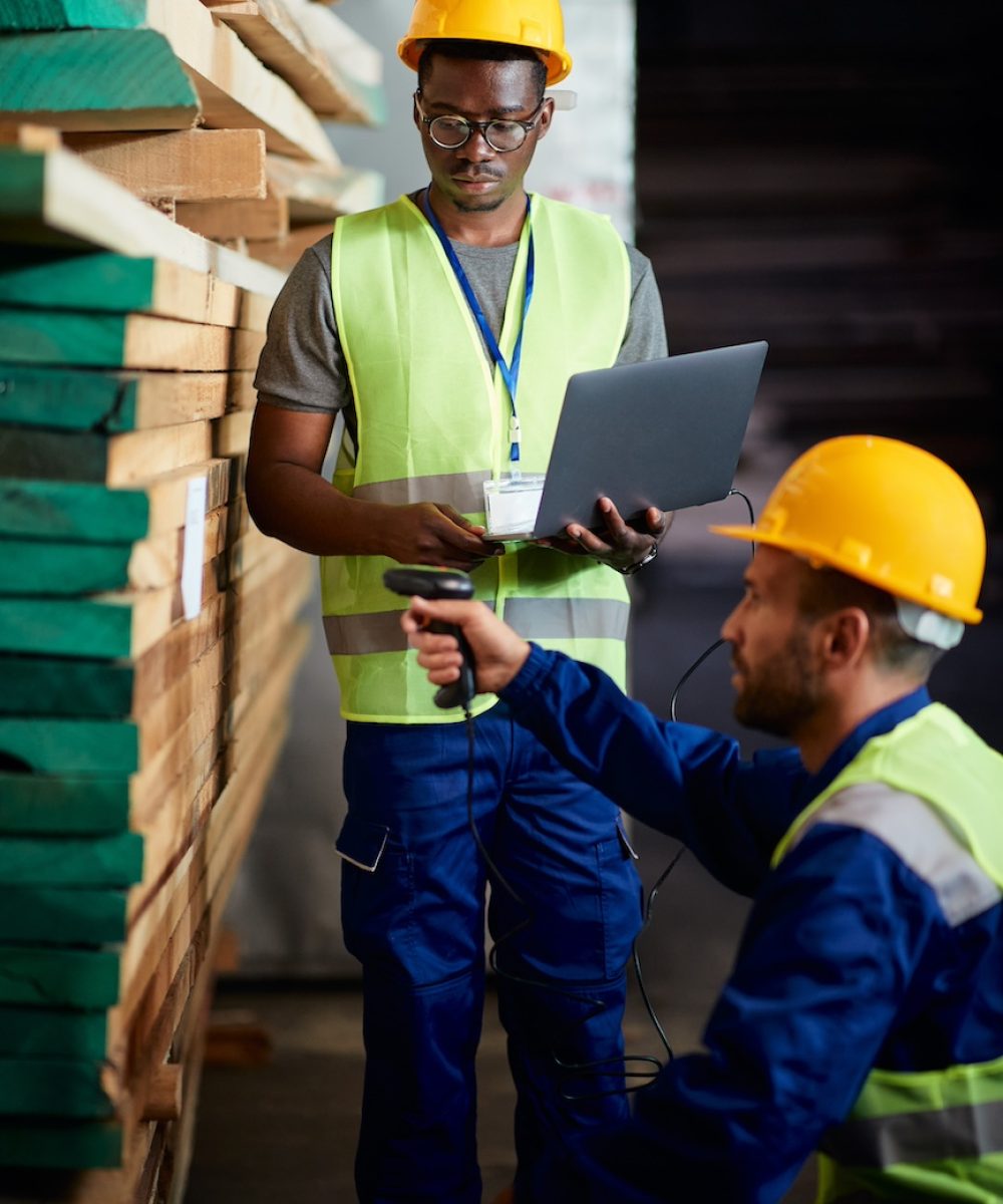 African American wood warehouse worker using laptop while his coworker is scanning labels at storage compartment.